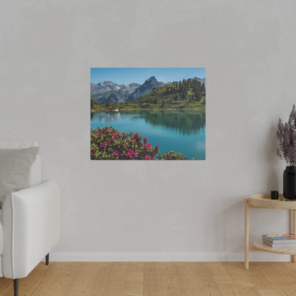 Square wall art of a blue alpine lake with mountains and pink flowers in the foreground, hung on a light gray wall.