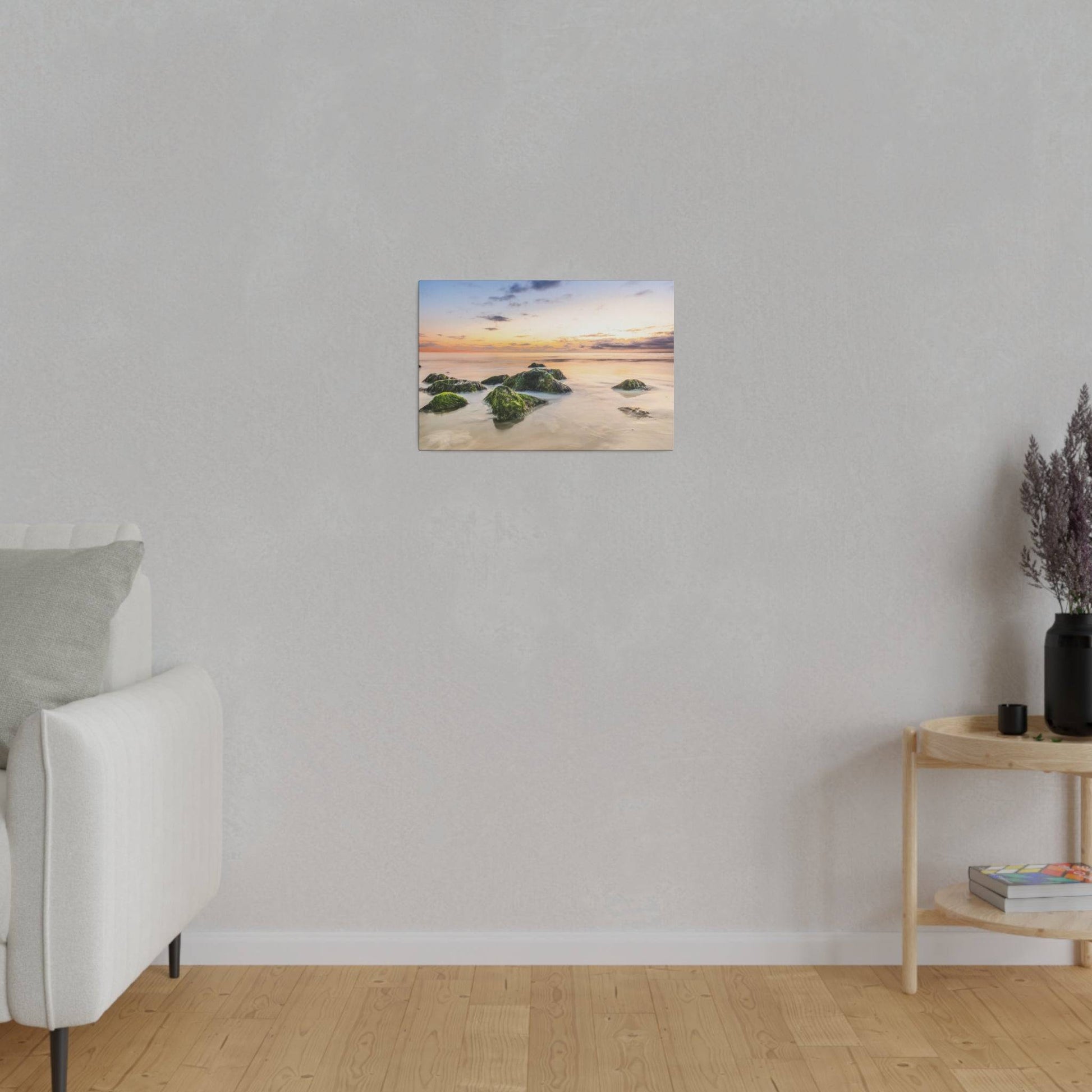 Small framed landscape painting of a rocky shore at sunset hangs on a light gray wall above a wooden floor.