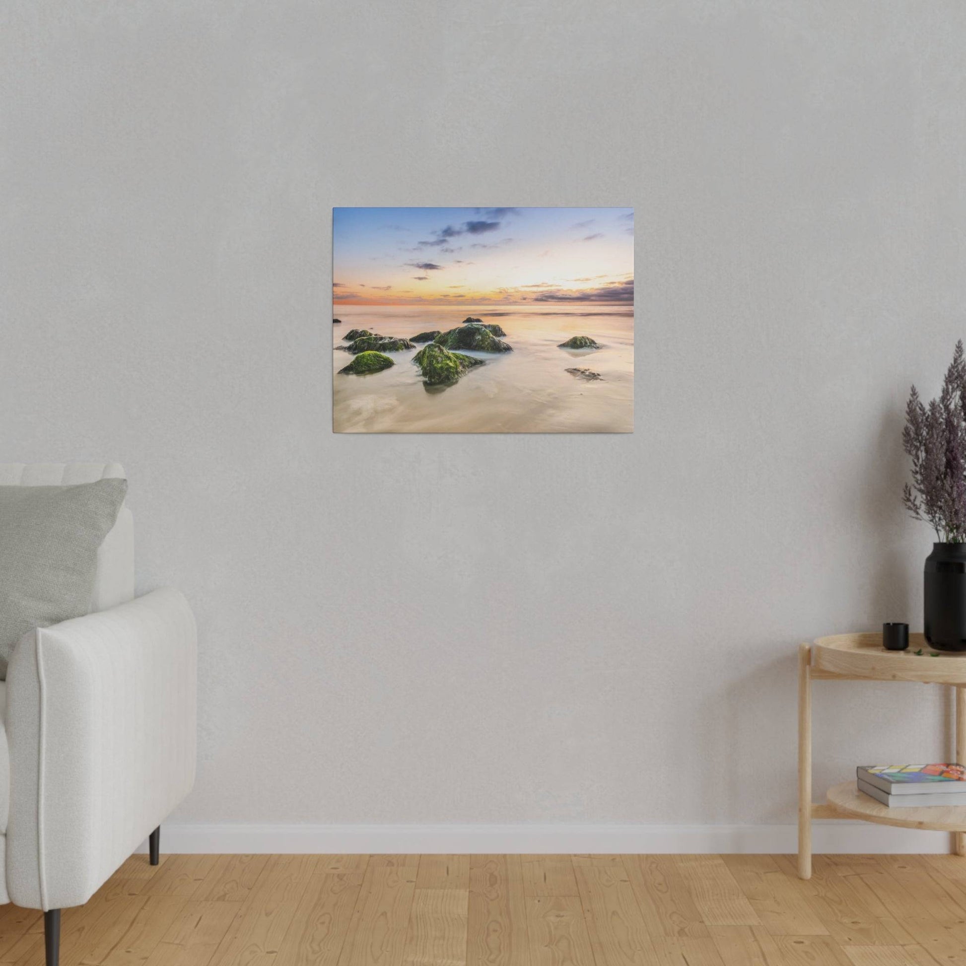 A coastal landscape painting of rocks in calm water at sunset hangs on a light gray wall above a wooden floor.