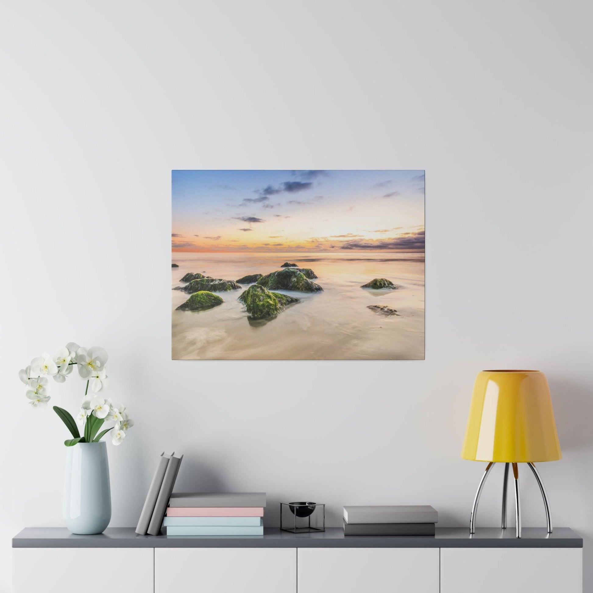 A square painting of a sunset over a rocky beach with calm water and mossy green rocks.