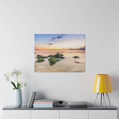 A square painting of a sunset over a rocky beach with calm water and mossy green rocks.