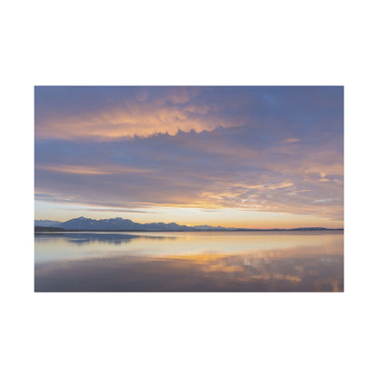Calm lake at dusk reflecting orange, pink, and purple sky with distant snow-capped mountains on the horizon.