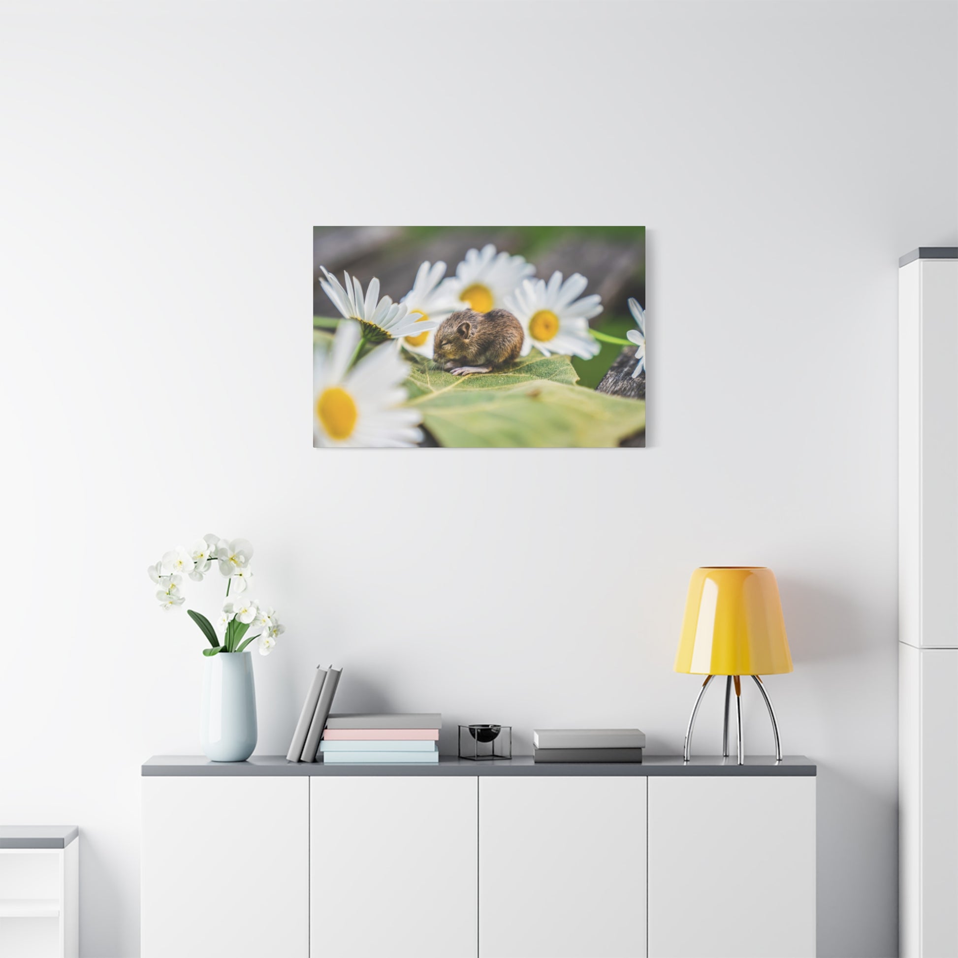 Framed photo of a small brown mouse on green leaves among white daisies, hung on a white wall above a cabinet.