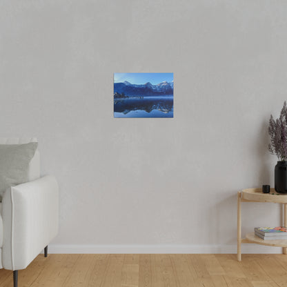 Small blue-toned landscape painting of a mountain lake reflected in calm water, hung on a light gray wall.