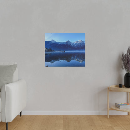 Square photo of blue mountains reflected in a calm lake, with a small pier on the left.
