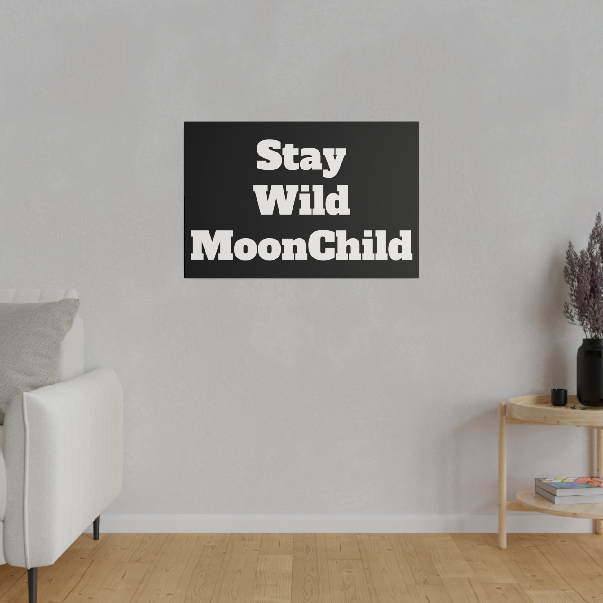 Black rectangular wall art with bold white text "Stay Wild MoonChild" mounted on a light gray living room wall.