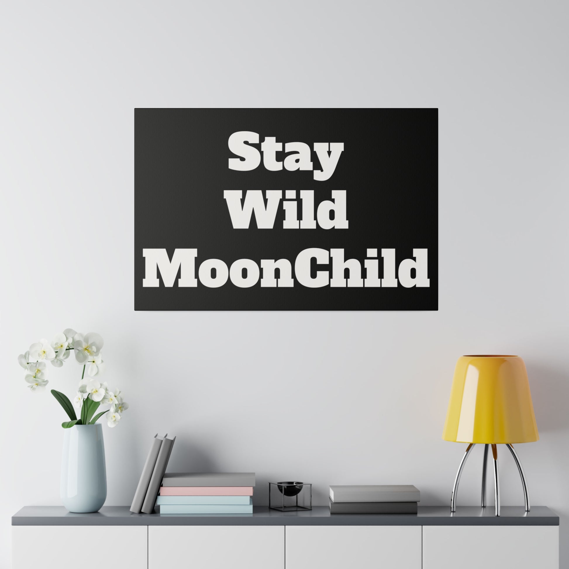 Black rectangular wall art reading 'Stay Wild MoonChild' in white typography, hung above a cabinet with a vase, books, and a yellow lamp.