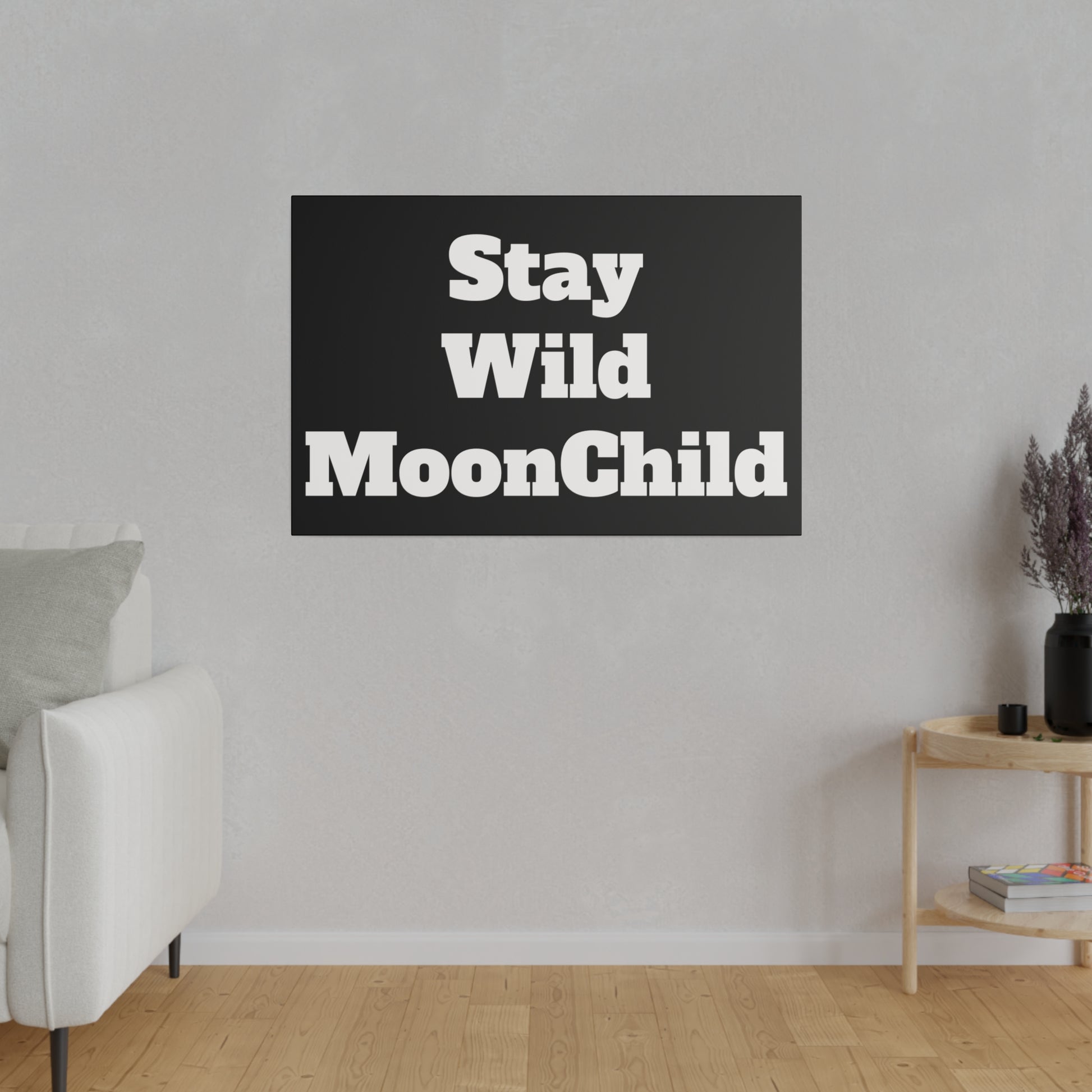 Black rectangular wall art with bold white text "Stay Wild MoonChild" hangs on a gray wall above a white sofa in a minimalist living room.