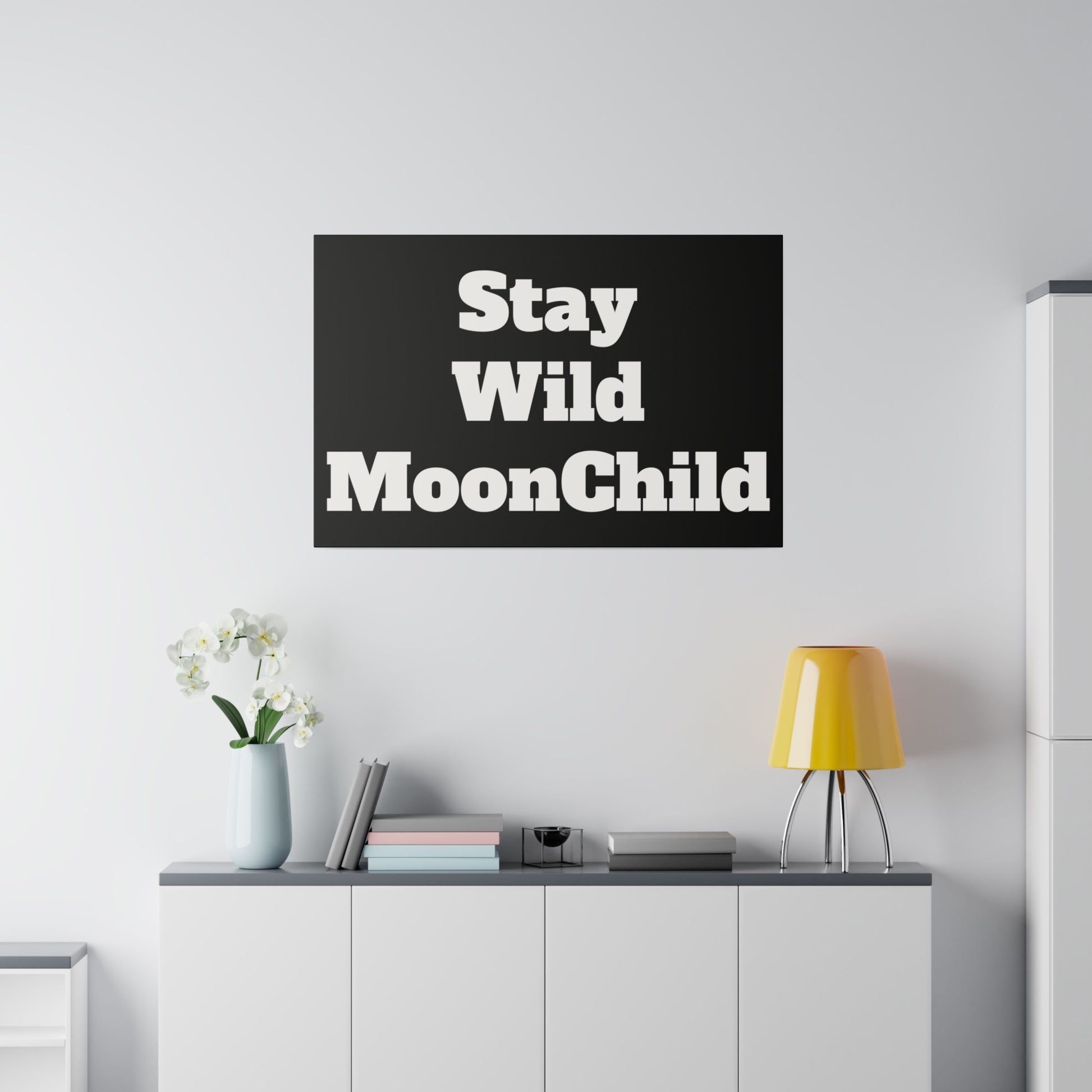 Black poster with bold white text “Stay Wild MoonChild” hangs on a white wall above a white cabinet with a vase, books, and a yellow lamp.