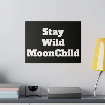 Black rectangular sign with bold white text reading “Stay Wild MoonChild” mounted on a light gray wall above a shelf.