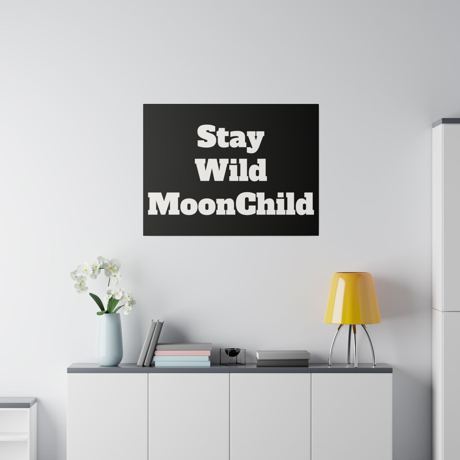 Black square wall art with bold white text "Stay Wild MoonChild" hung above a white sideboard in a minimalist living room.