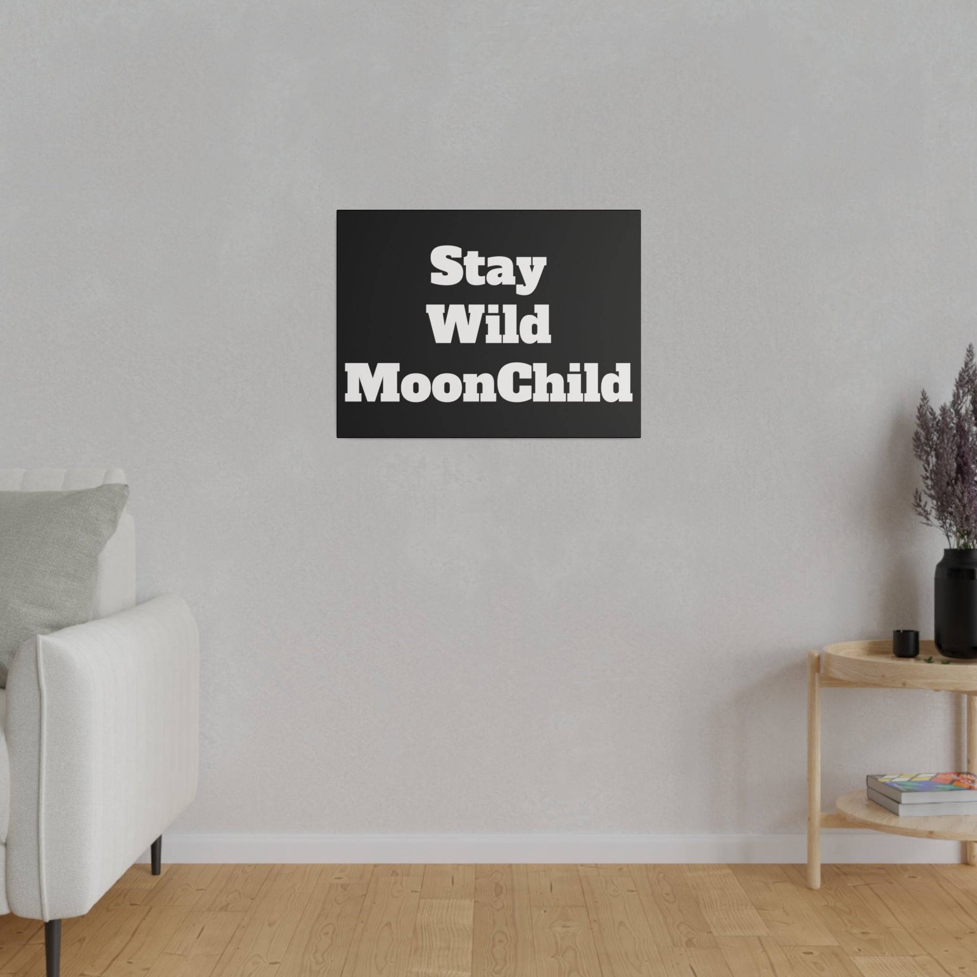 A black rectangular wall art with bold white text 'Stay Wild MoonChild' hangs on a light gray wall above a wooden floor.