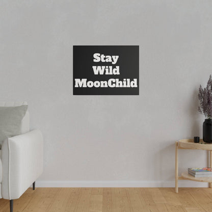 A black rectangular wall art with bold white text 'Stay Wild MoonChild' hangs on a light gray wall above a wooden floor.