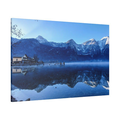 Blue alpine lake scene with snow-capped mountains, a wooden pier, and a small house along the shore reflected in calm water.