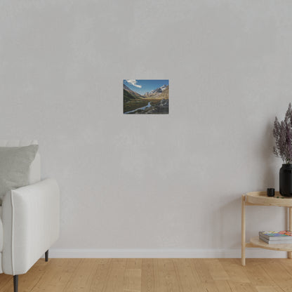 Small landscape painting of a mountain valley with a river, hung on a light gray wall above a wooden floor.