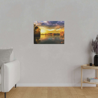 Framed landscape painting of a sunset over a calm river with trees along the banks, hung on a light gray wall.