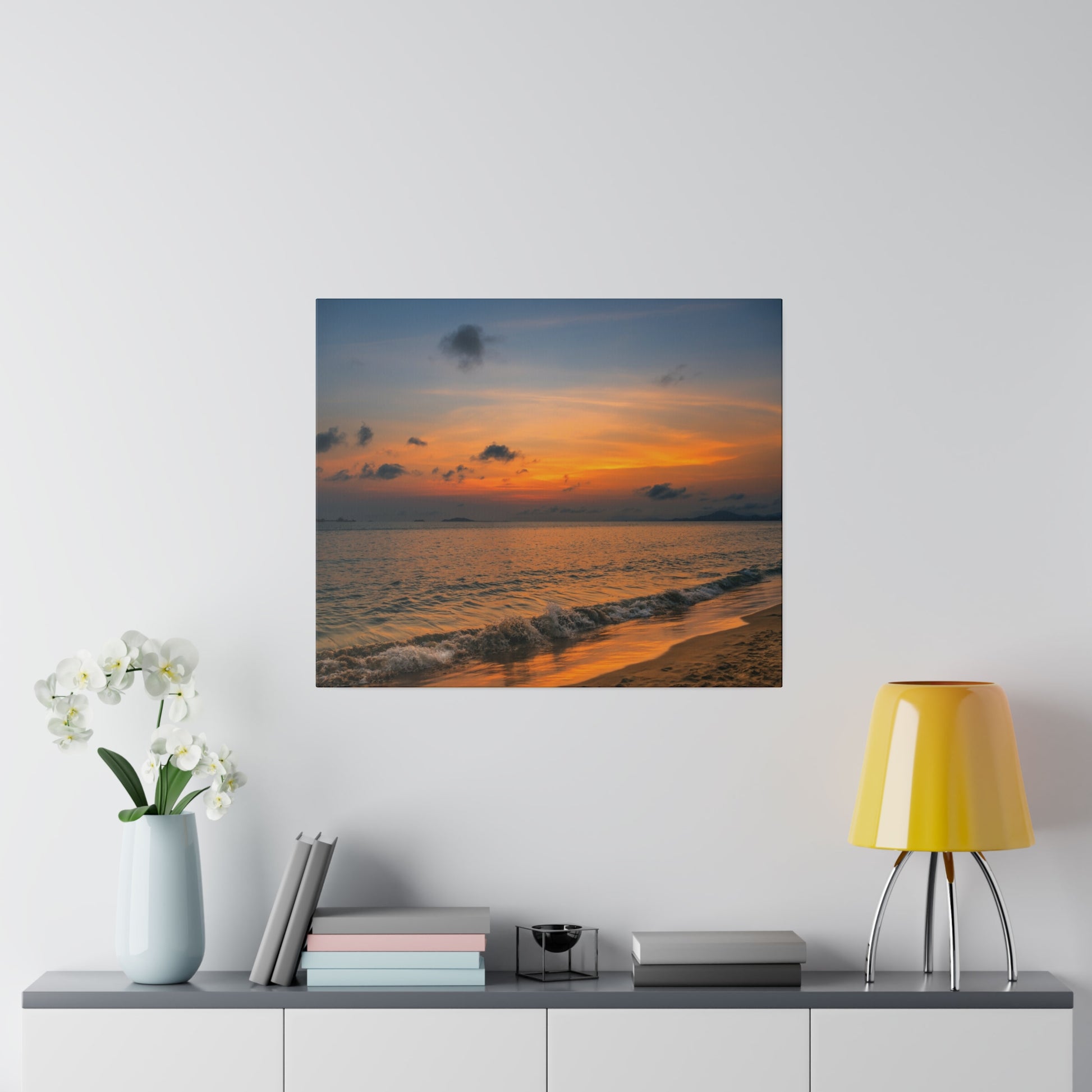 Square wall art showing a sunset over the ocean with orange sky, dark clouds, and gentle waves on the sandy shore.