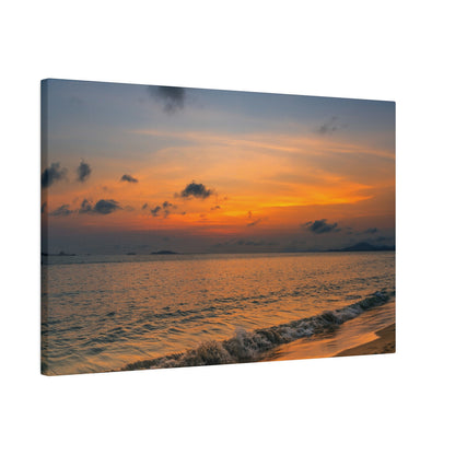 Photo of a calm ocean at sunset with an orange-yellow sky and small waves washing onto a sandy shore.