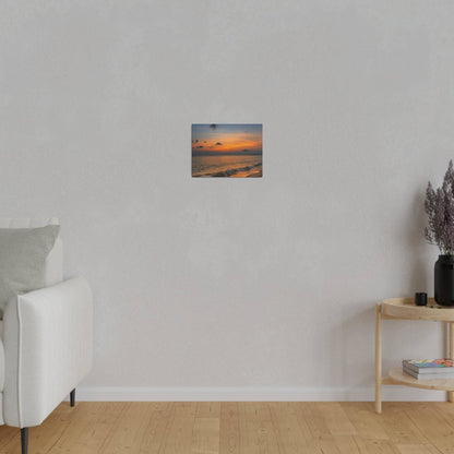Small square sunset photograph hung on a light gray wall above a wooden floor, with a white sofa to the left and a side table on the right.