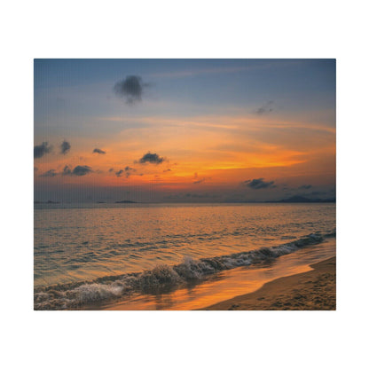 Sunset over the ocean with orange and blue sky, small clouds, and gentle waves washing onto a sandy beach.