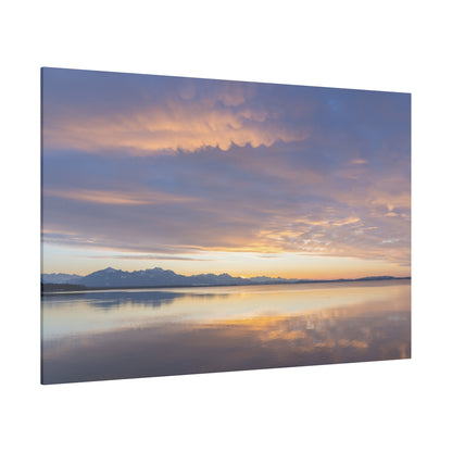 Calm lake with distant snow-capped mountains under a sunset sky, reflecting orange, pink and blue hues.