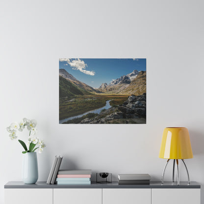 Photograph of a mountain valley with a winding river, rocky foreground, green slopes, and snow-capped peaks under a clear blue sky.