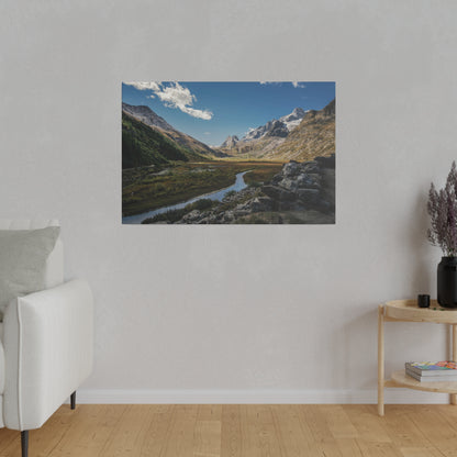 Framed landscape photo of a mountain valley with a winding river, rocky foreground, and snow-capped peaks under a blue sky.