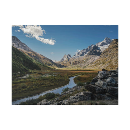 Rugged mountain valley with a winding river, grassy meadows, and a rocky foreground under a blue sky.