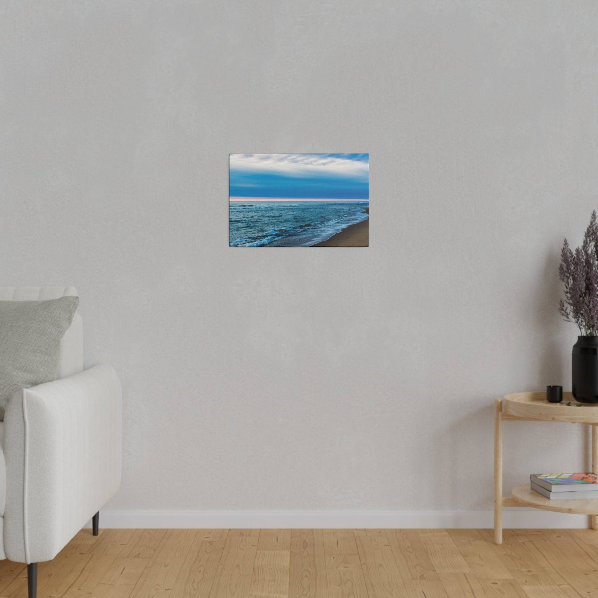 Small coastal painting of a blue ocean and beach hangs on a light gray wall above a white sofa.