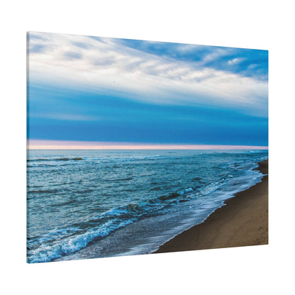 Photo of a sandy beach with blue ocean waves gently washing ashore under a cloudy blue sky.