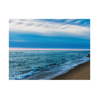 A seascape showing blue ocean waves lapping a sandy beach under a pale pink and blue sky.