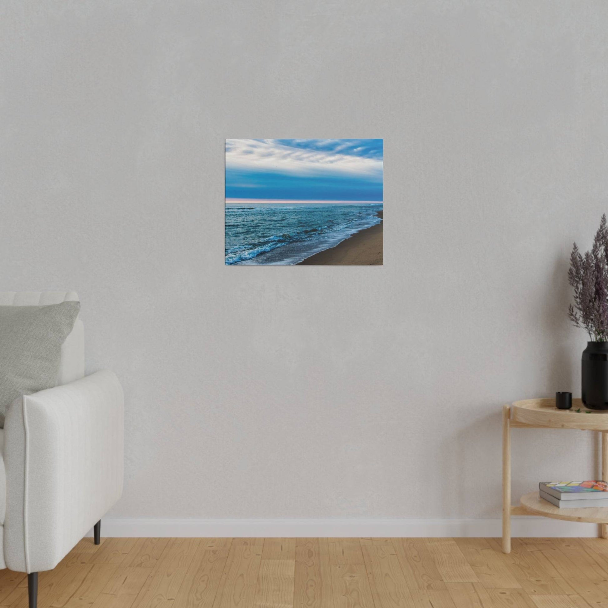 Small square painting of a blue ocean and sandy beach hangs on a light gray wall above a wooden floor.