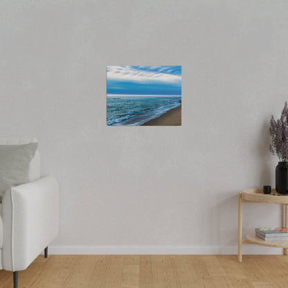 Small square painting of a blue ocean and sandy beach hangs on a light gray wall above a wooden floor.