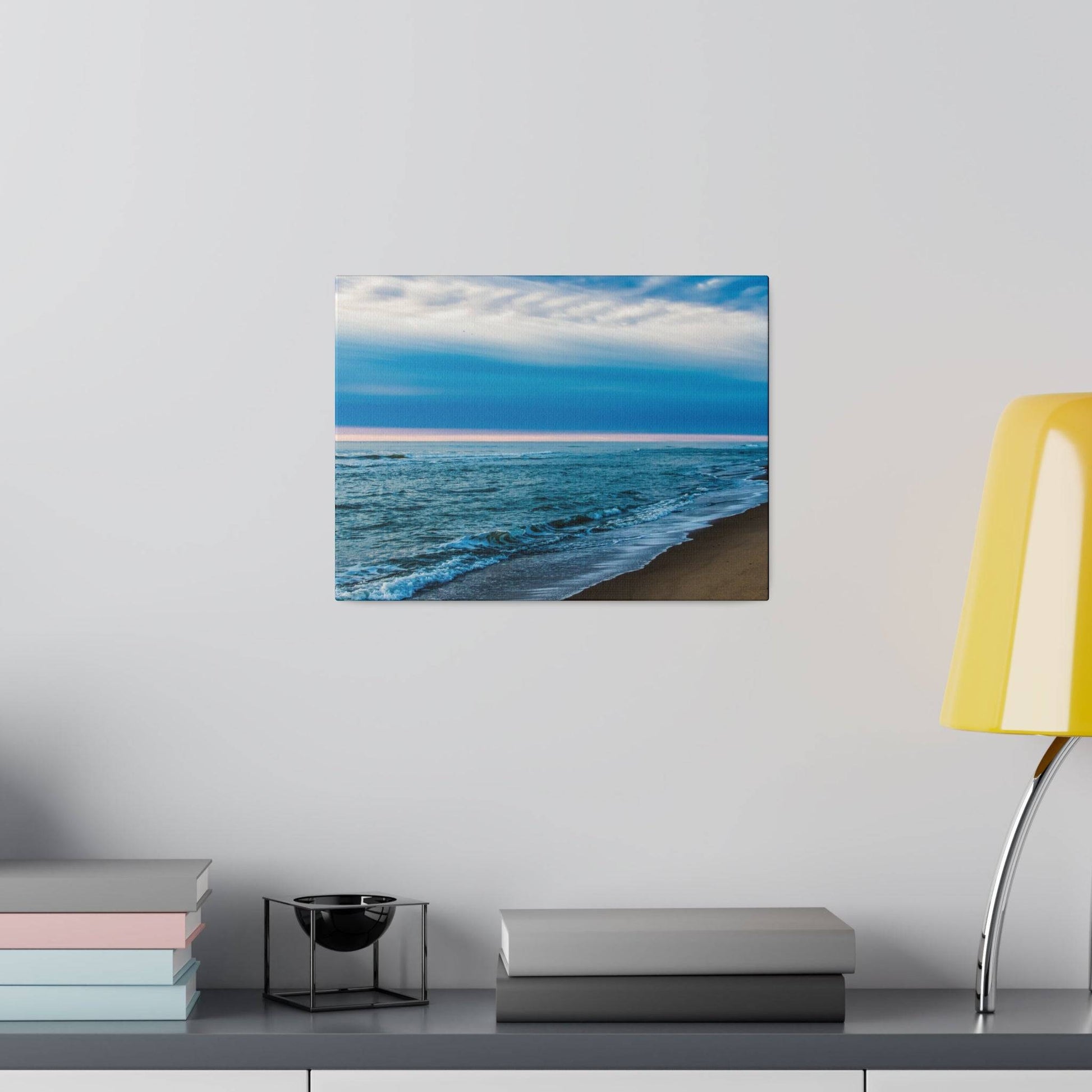 Square wall art showing a blue ocean shoreline with waves crashing onto a sandy beach under a cloudy sky.