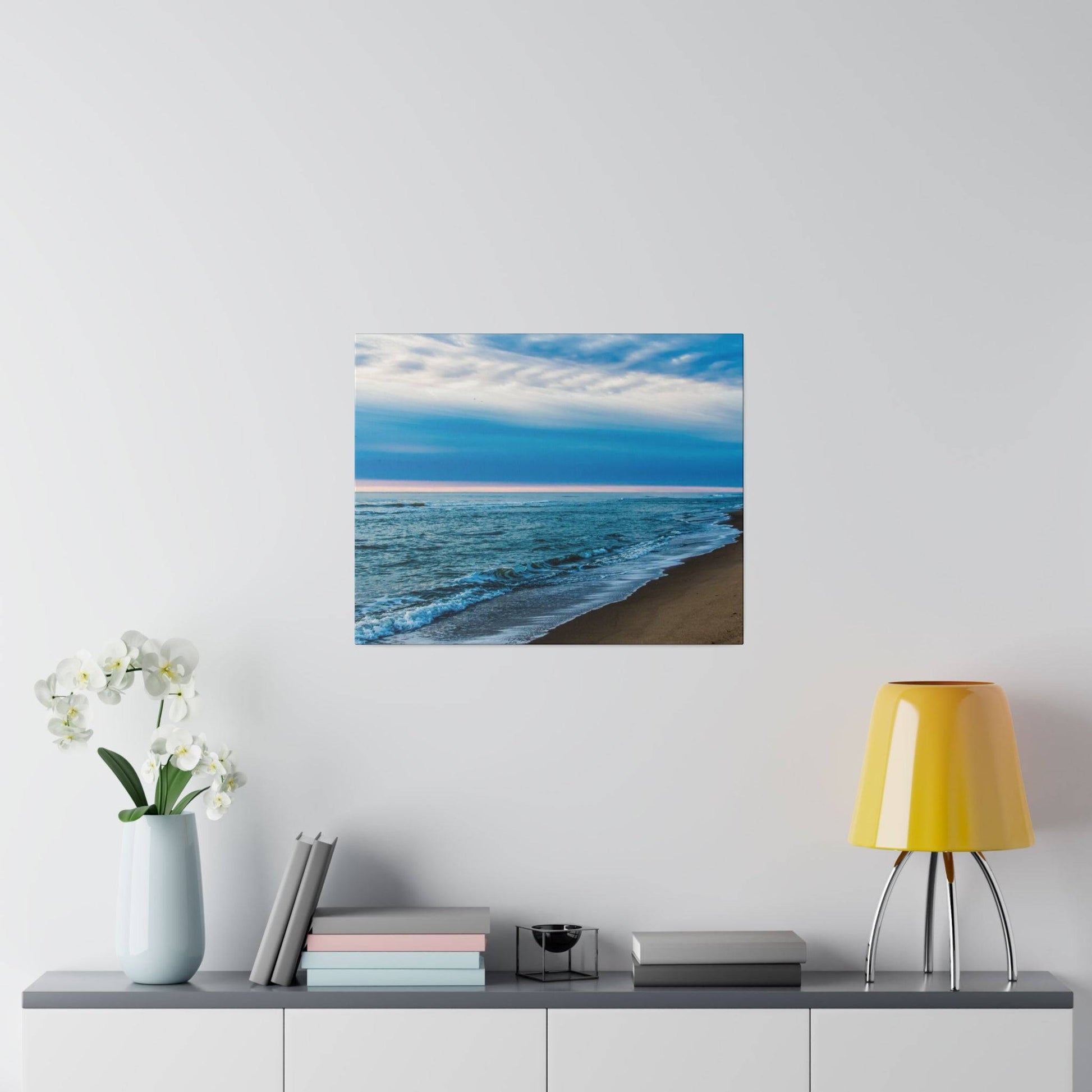 Square print of a blue ocean beach with waves along a sandy shore under a cloudy sky, mounted on a light gray wall.