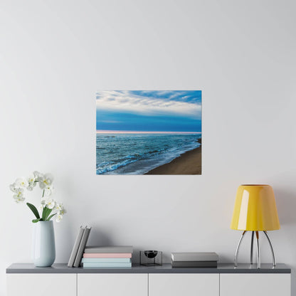 Square print of a blue ocean beach with waves along a sandy shore under a cloudy sky, mounted on a light gray wall.