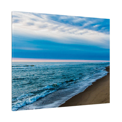 Image of a sandy beach with blue ocean waves and white foam under a blue, cloudy sky.