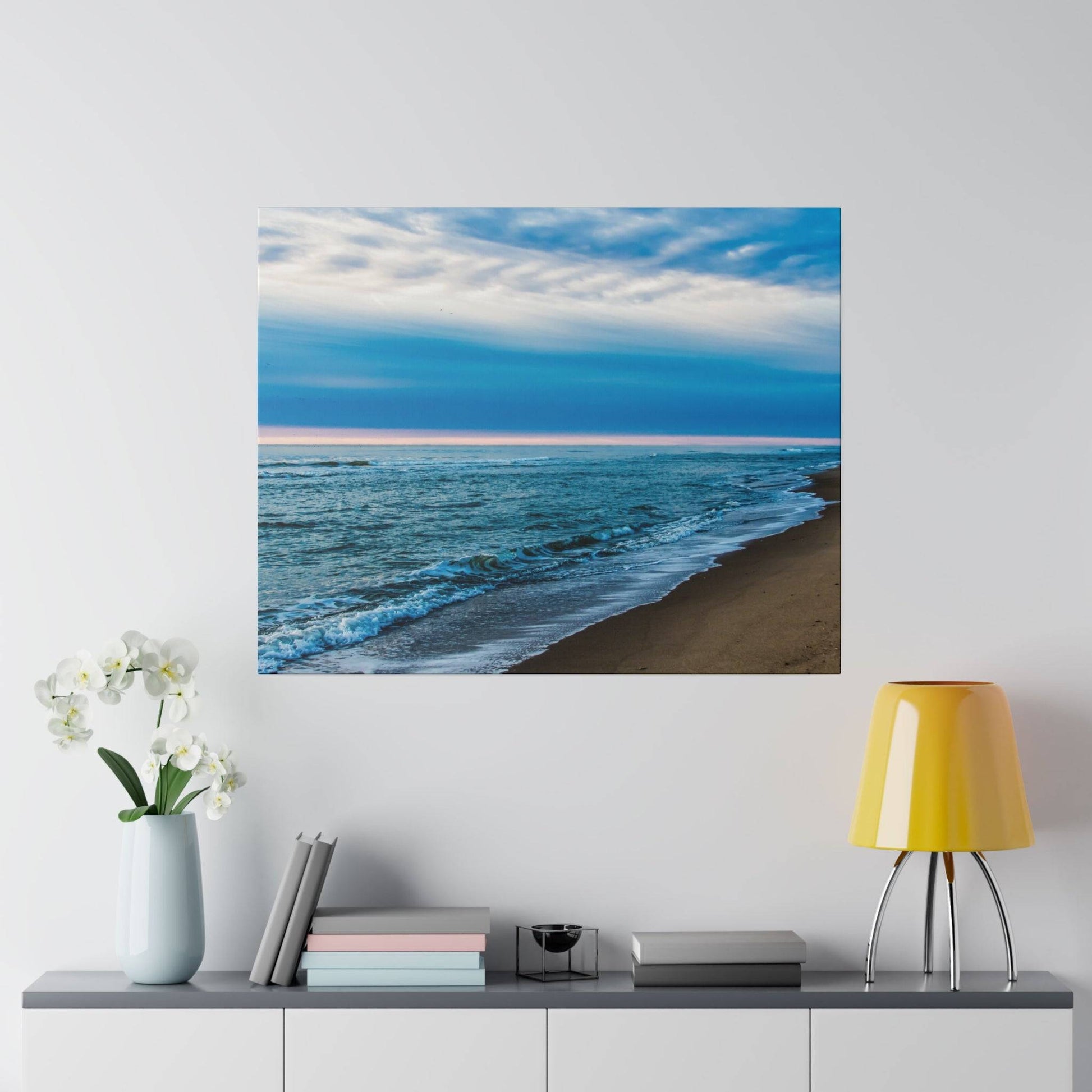 Square wall art showing a blue ocean beach with white waves along a sandy shore under a cloudy sky.