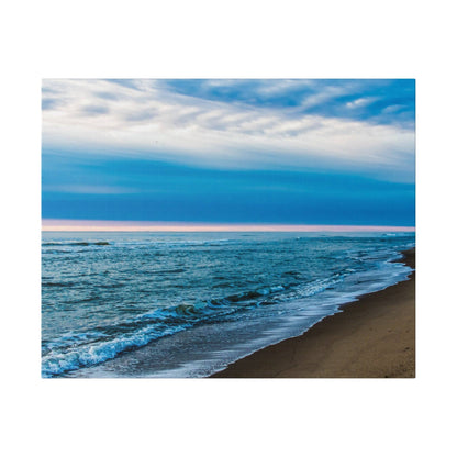 A beach scene with blue ocean waves washing onto brown sand under a blue sky with wispy clouds.