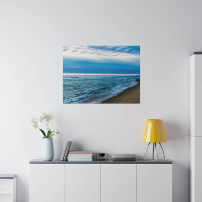 Seascape painting of a blue ocean and sandy shore hangs above a white sideboard with a yellow lamp and vase of white orchids.