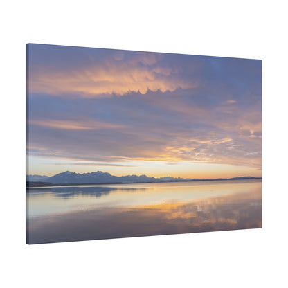 Calm lake at sunset reflecting orange and purple sky with distant snow-capped mountains.
