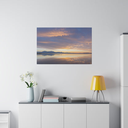 Wall art depicting a sunset over a calm lake with distant mountains and orange-purple clouds reflecting on the water.
