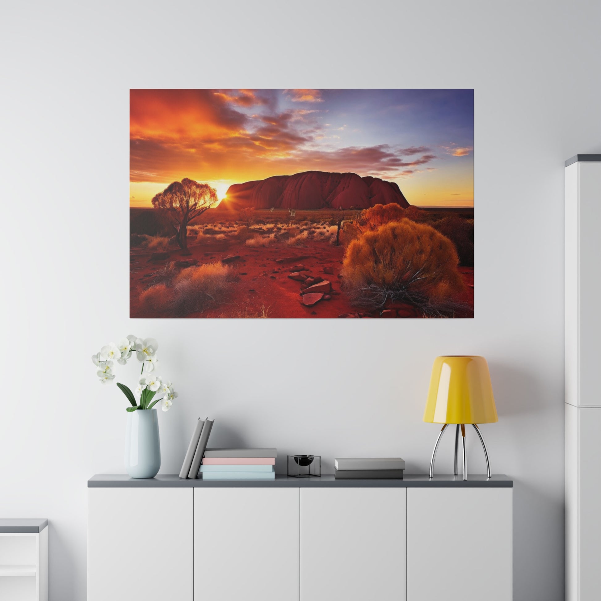 Large wall art depicting Uluru at sunset over a red desert, hung above a white sideboard with a yellow lamp.