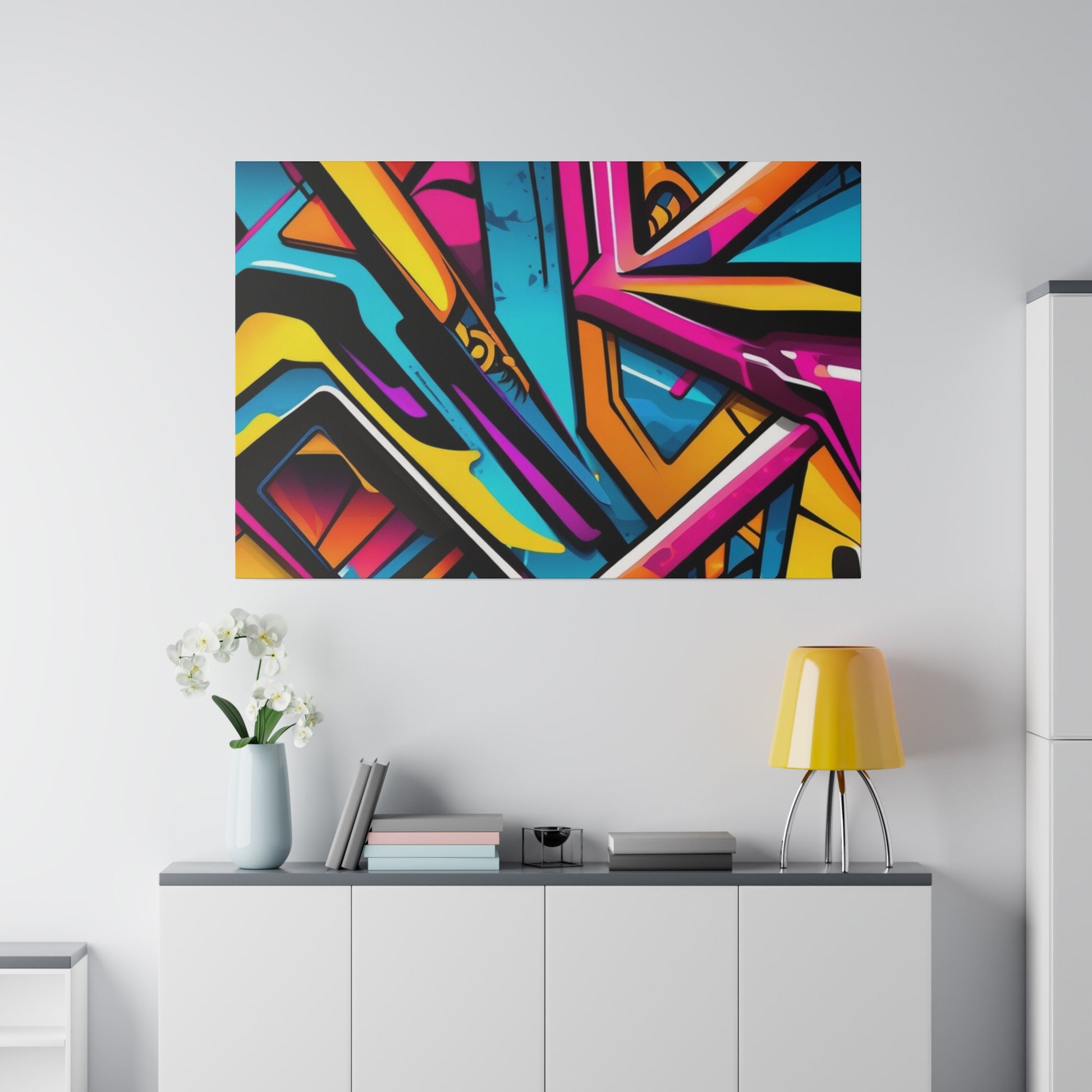 Colorful abstract graffiti art mounted on a light gray wall above a white console with books, a vase, and a yellow lamp.