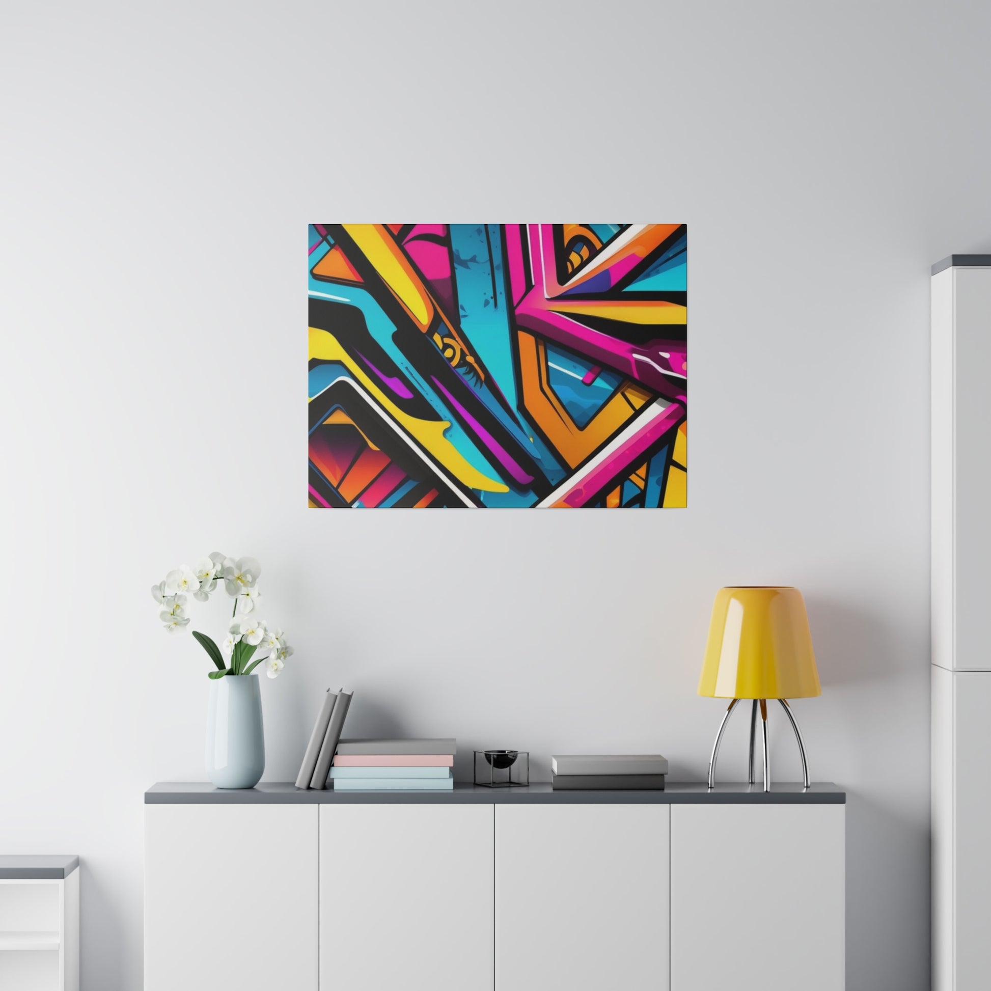 Colorful abstract graffiti artwork on a white wall above a white cabinet with a yellow lamp and a vase.