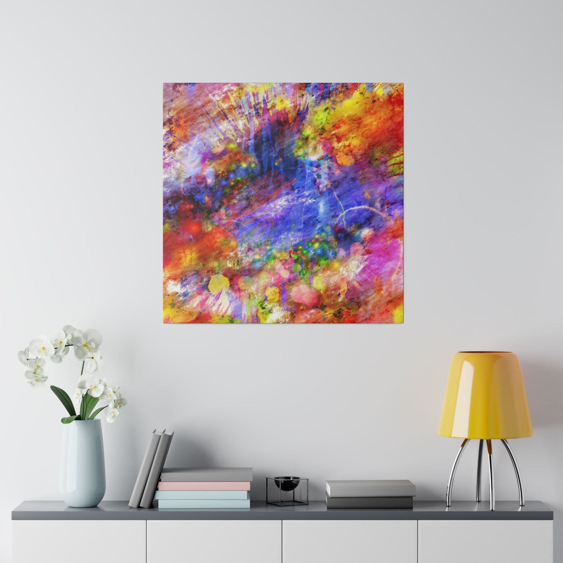 Square abstract painting in vivid reds, blues, and yellows hanging above a gray console with a pale blue vase of white orchids, books, and