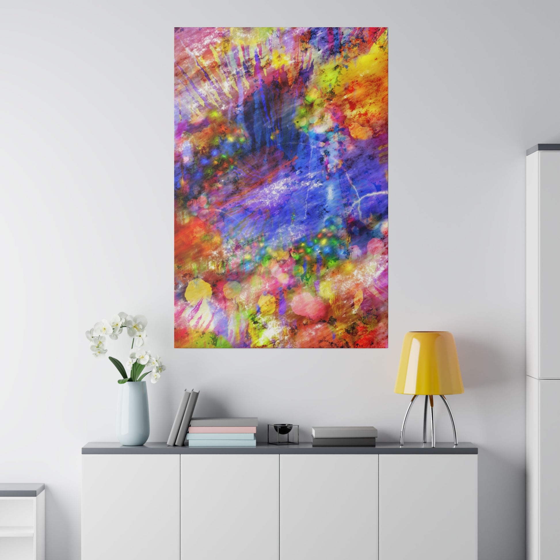 Vertical abstract painting with bright multicolor splashes hangs above a white cabinet, beside a pale blue vase of white flowers and a