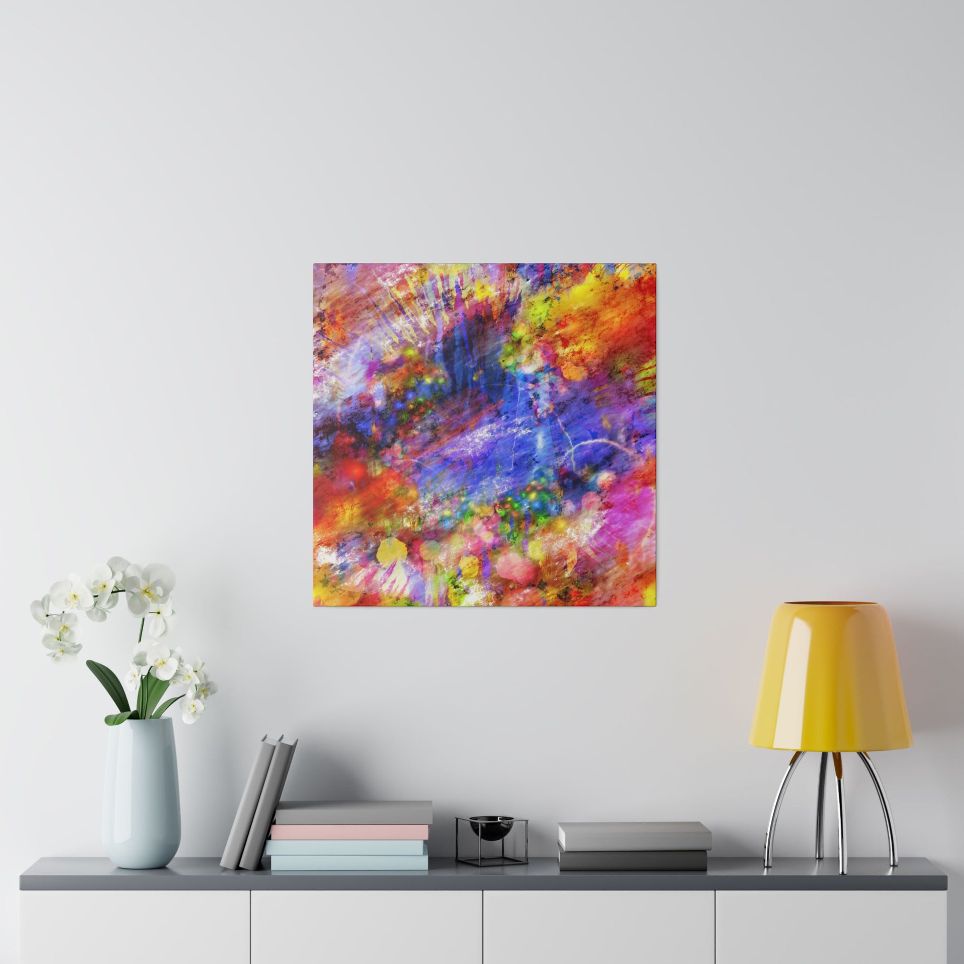 Abstract multicolored painting hung on a white wall above a gray console with a blue vase of white flowers, books, and a yellow lamp.