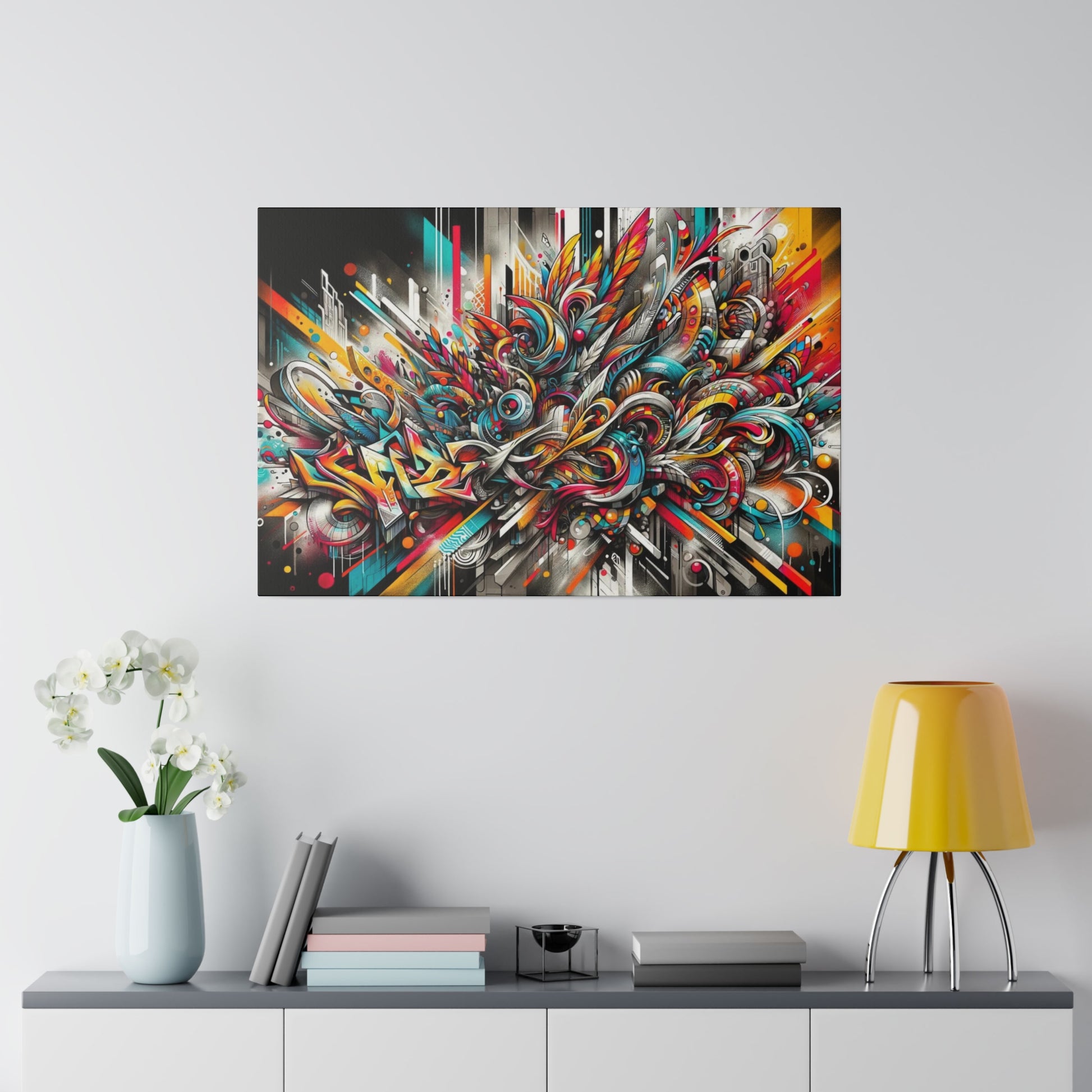 Colorful abstract graffiti-style painting on canvas mounted on a light gray wall above a sideboard with a vase, books, and a yellow lamp.