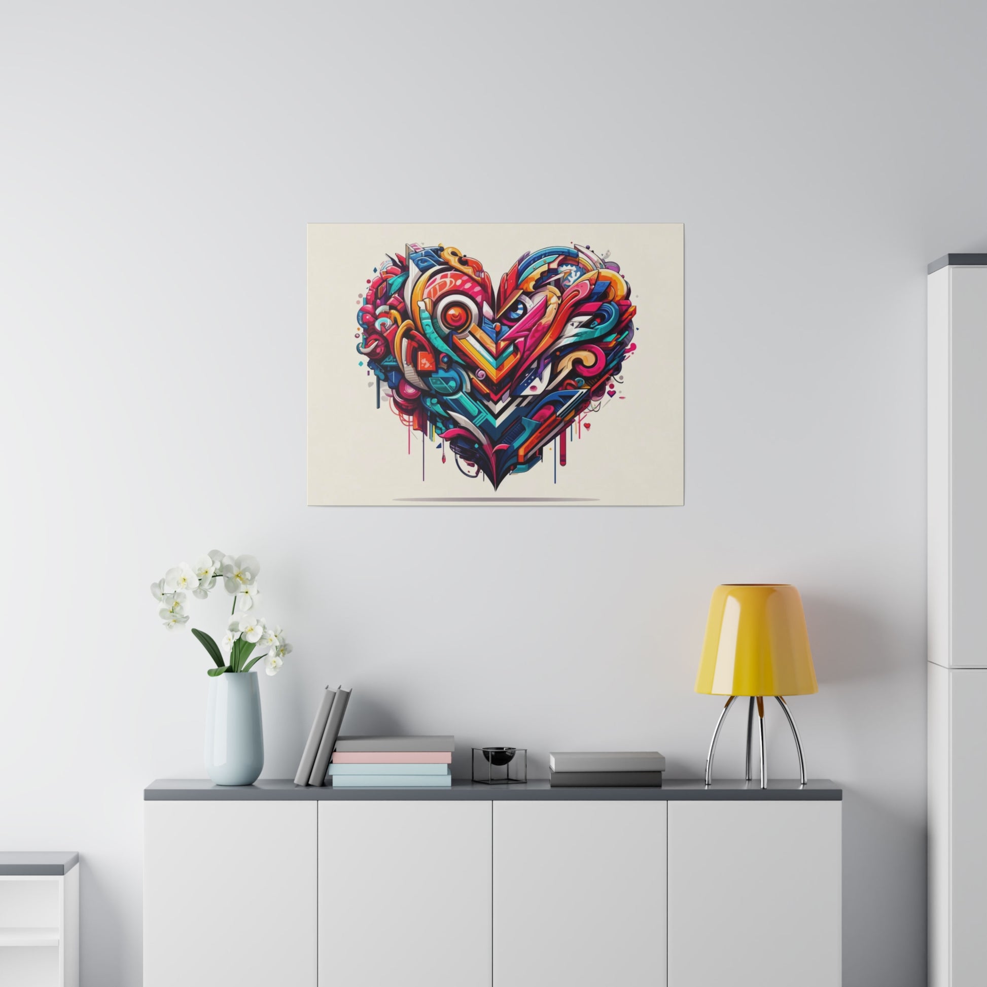 Colorful abstract heart graffiti artwork mounted on a white wall above a white cabinet with a yellow lamp and white flowers.